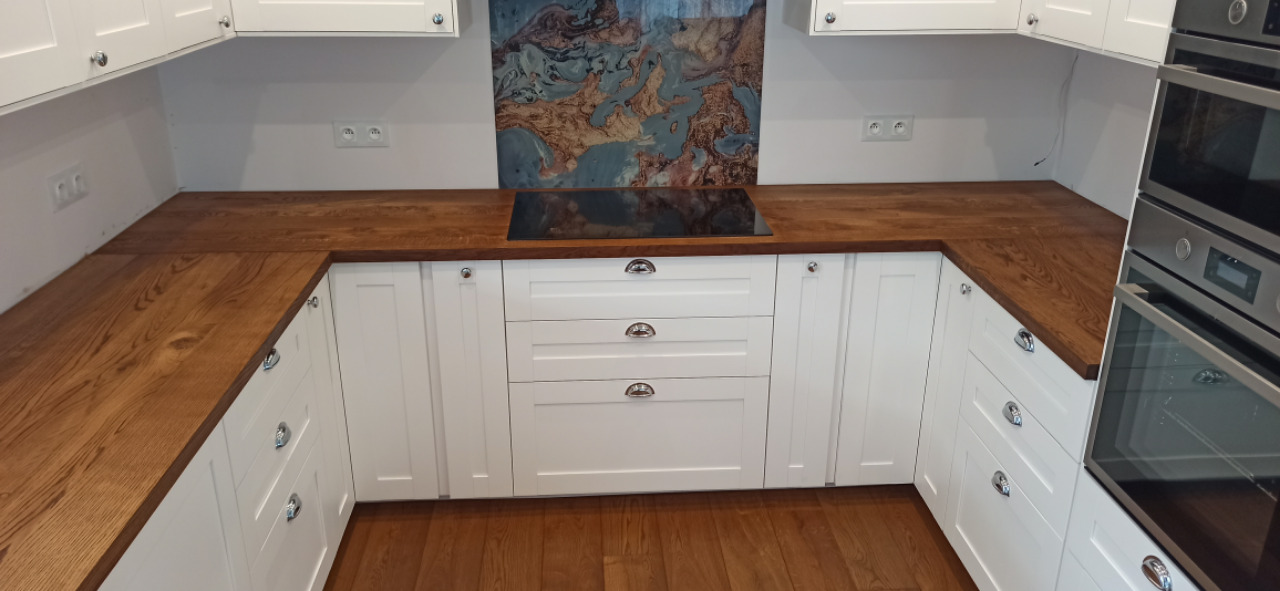 Kuchenny blat drewniany o ciepłym odcieniu, zamontowany w białej zabudowie meblowej w Łagiewnikach Nowych. Widoczna płyta indukcyjna i piekarnik w zabudowie.