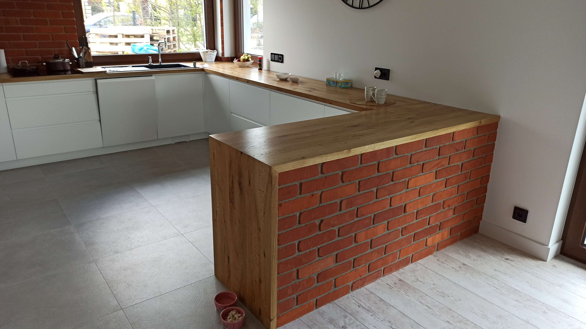 Nowoczesna kuchnia z drewnianym blatem i wyspą wykończoną czerwoną cegłą, białe szafki, jasna podłoga, widoczne naczynia i owoce na blacie.
