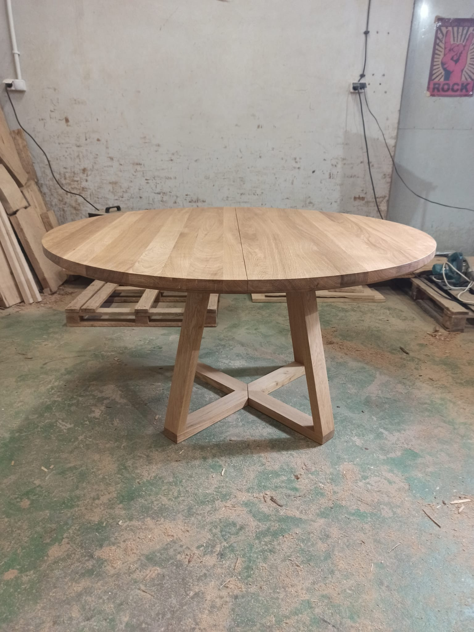 Okrągły, rozkładany stół z jasnego drewna z charakterystyczną podstawą w kształcie litery X, widoczny w warsztacie z surowym tłem i plakatem 'Rock'.