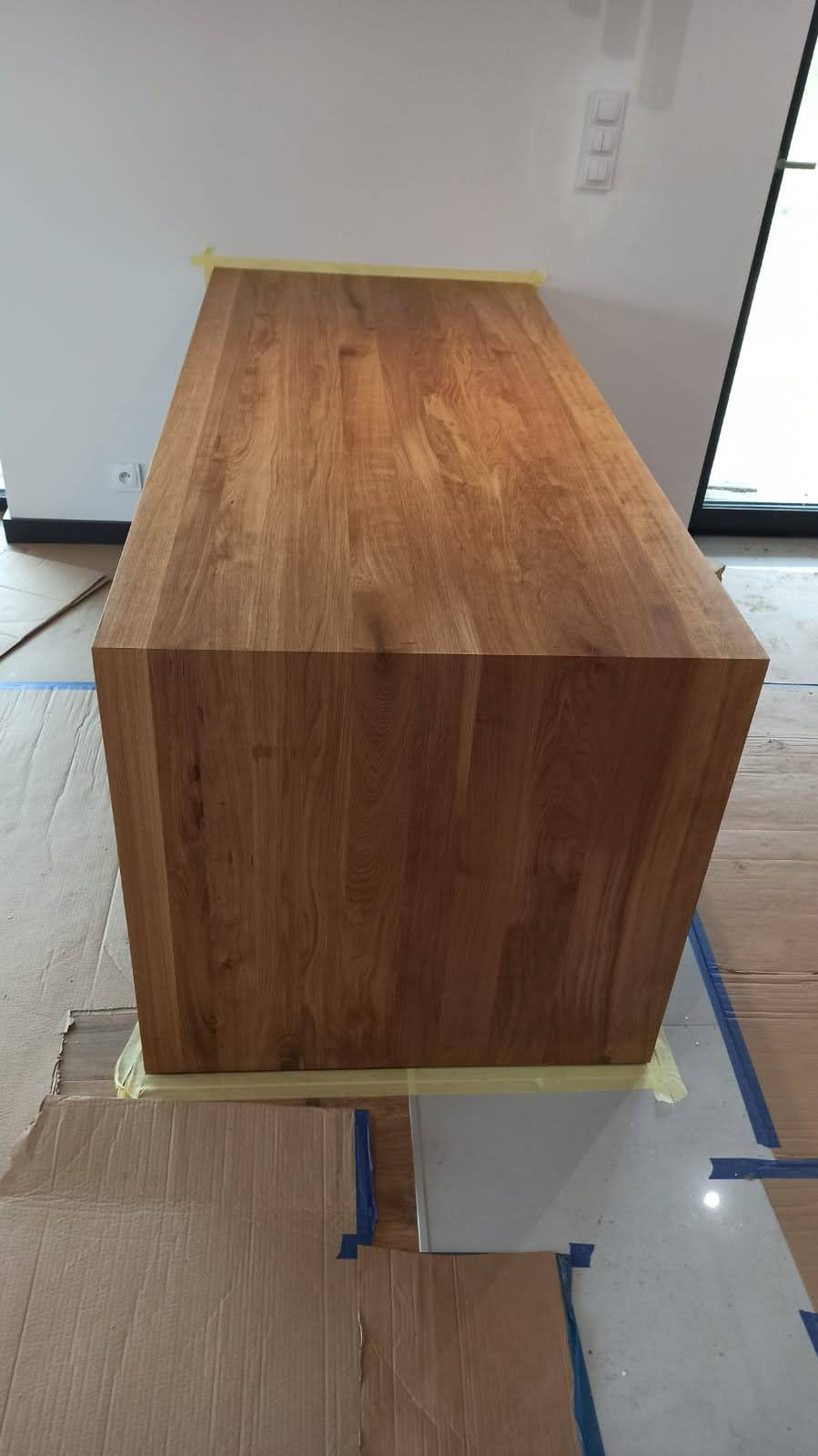 Blat drewniany z litego dębu, zabezpieczony taśmą malarską na czas prac wykończeniowych w pomieszczeniu z jasnymi ścianami i dużym oknem.