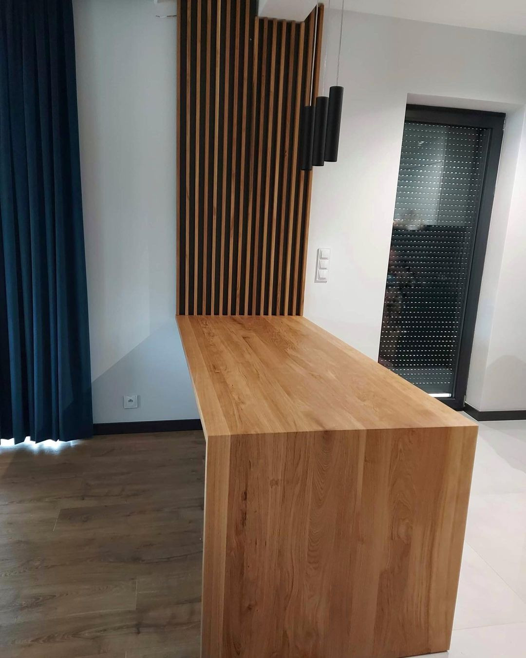 Jasny, dębowy blat kuchenny z litego drewna, połączony z pionową, drewnianą lamelową ścianką dekoracyjną, oświetlony trzema czarnymi lampami wiszącymi, na tle zasłoniętego okna i fragmentu zasłony...
