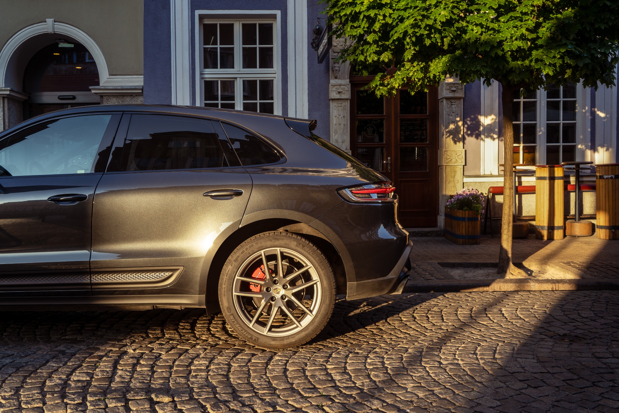 Szary Porsche Macan zaparkowany na brukowanej ulicy w Gdańsku, z widocznym fragmentem budynku w tle i cieniem drzewa padającym na bruk.