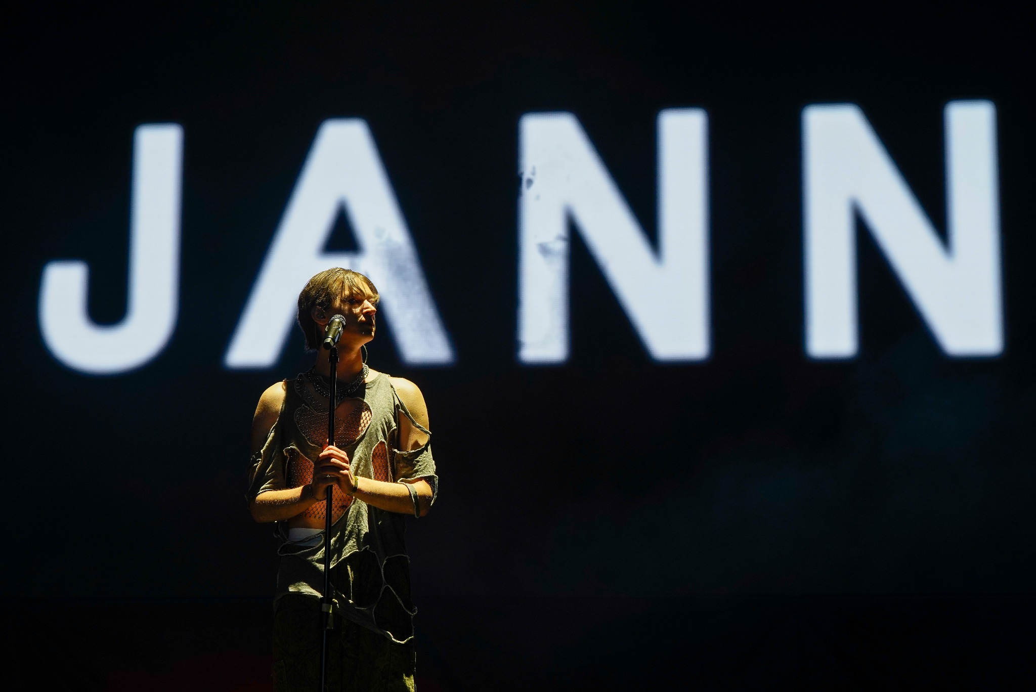 Piosenkarz Jann w podziurawionej koszulce śpiewa do mikrofonu na scenie Opener 2023, w tle napis z jego imieniem.