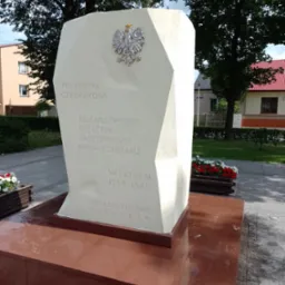 Pomnik z jasnego kamienia upamiętniający poległych członków ruchu oporu przeciwko faszyzmowi niemieckiemu w latach 1939-1945, z godłem Polski, umieszczony na cokole z czerwonego kamienia, otoczony...