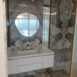 Elegancka łazienka z umywalką nablatową i okrągłym lustrem na tle ściany wykończonej płytami imitującymi marmur z wyraźnym użyleniem, złota armatura i białe szafki z ryflowanymi frontami.