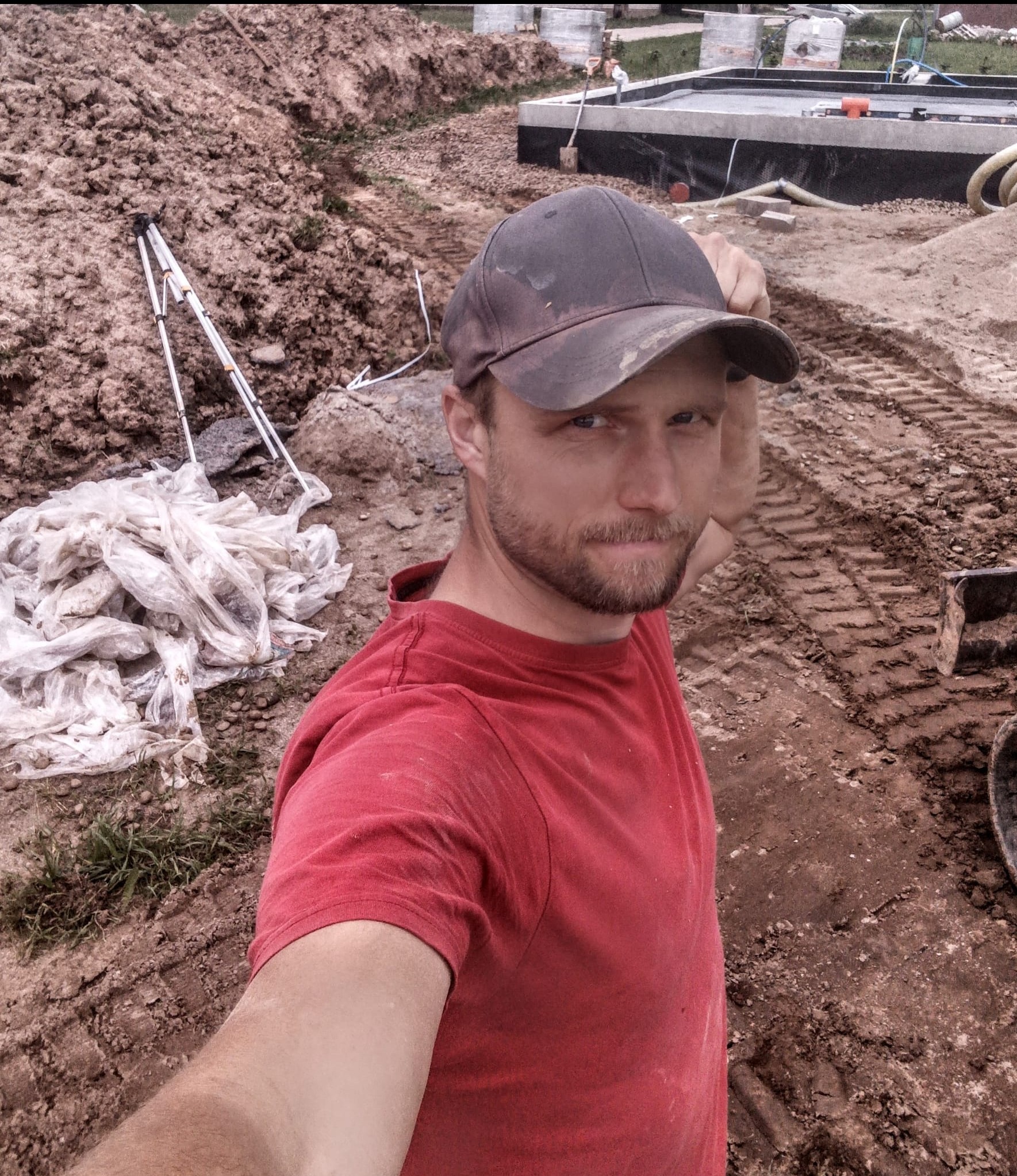 Fundament domu w trakcie budowy, widoczny z perspektywy osoby robiącej selfie. W tle ziemia, narzędzia i materiały budowlane. Ujęcie z dołu, lekko amatorskie.