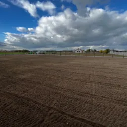 Przygotowany grunt pod trawnik, widoczne ślady po bronowaniu, w tle maszyny rolnicze i budynek w trakcie budowy pod błękitnym niebem z chmurami.