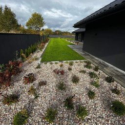 Garden - Nowoczesny ogród z równo przystrzyżonym trawnikiem, rabatą wysypaną drobnymi kamieniami i nasadzeniami ozdobnych krzewów oraz drzewek, obok ciemnego budynku z czarną dachówką i wysokiego, czarnego...