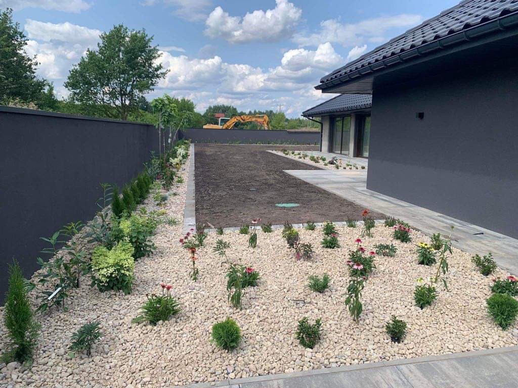 Nowo założony ogród z rabatą wysypaną drobnymi kamieniami, świeżo posadzone kwiaty i krzewy, przygotowany teren pod trawnik, nowoczesny dom w tle, widoczny fragment ogrodzenia i koparka w oddali.