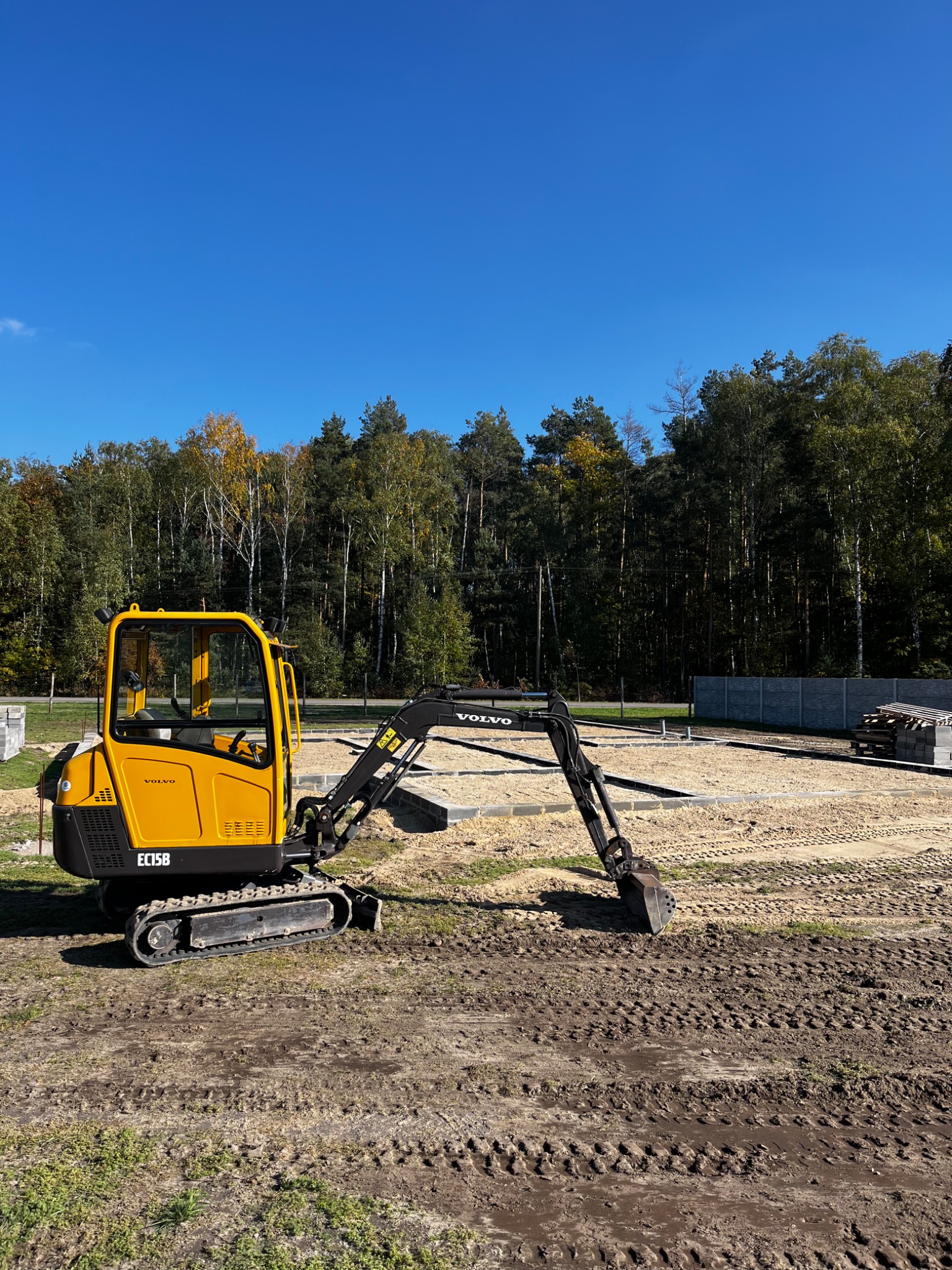 Żółta mini koparka Volvo EC15B na placu budowy, przygotowującym teren pod fundamenty, z widocznymi śladami gąsienic na ziemi i lasem w tle.