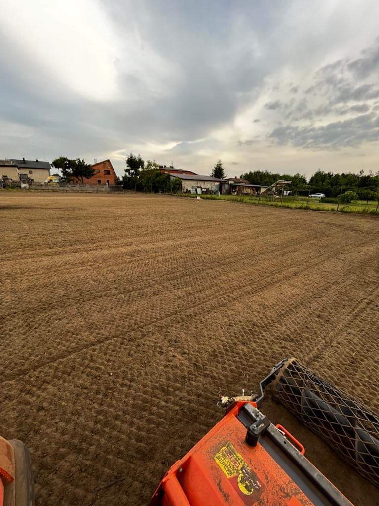 Przygotowane pole pod zasiew trawy, widoczne ślady po walcowaniu gleby, w tle zabudowania mieszkalne pod pochmurnym niebem.