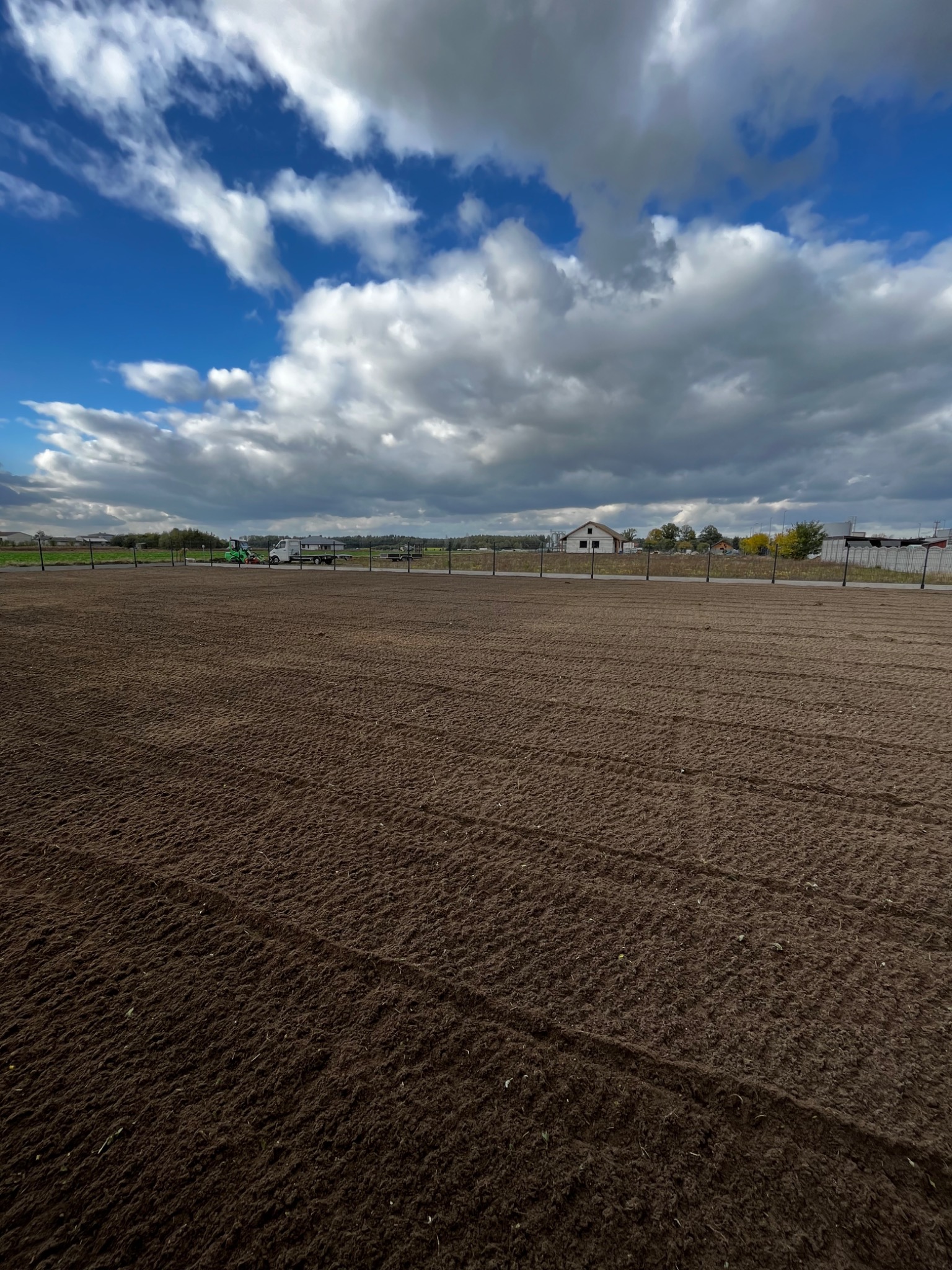 Przygotowany grunt pod trawnik, widoczne ślady po bronowaniu, w tle maszyny rolnicze i budynek w trakcie budowy pod błękitnym niebem z chmurami.