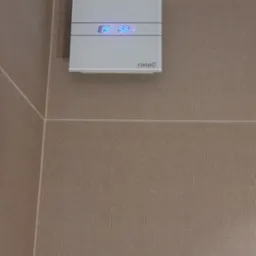 Biały wentylator łazienkowy z wyświetlaczem temperatury i wilgotności, zamontowany na beżowych płytkach ściennych w rogu pomieszczenia.