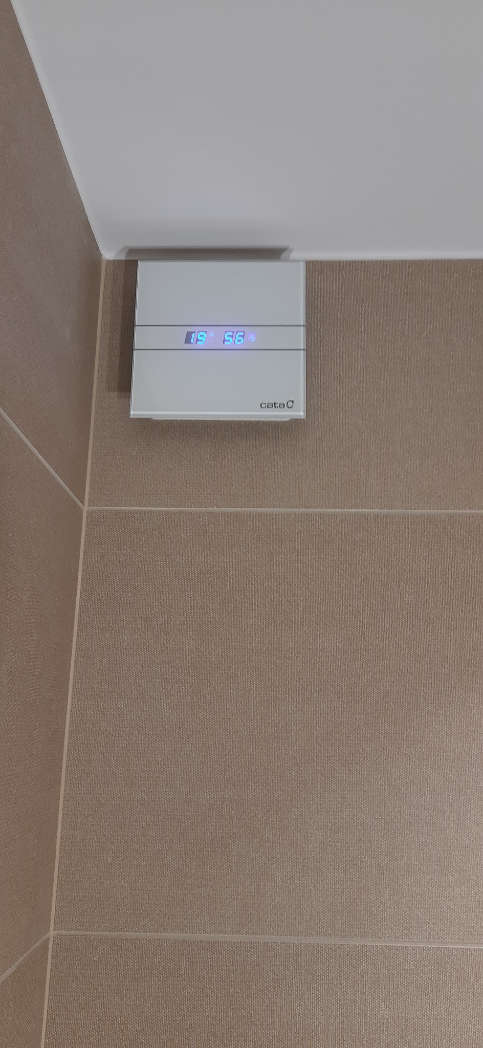 Biały wentylator łazienkowy z wyświetlaczem temperatury i wilgotności, zamontowany na beżowych płytkach ściennych w rogu pomieszczenia.