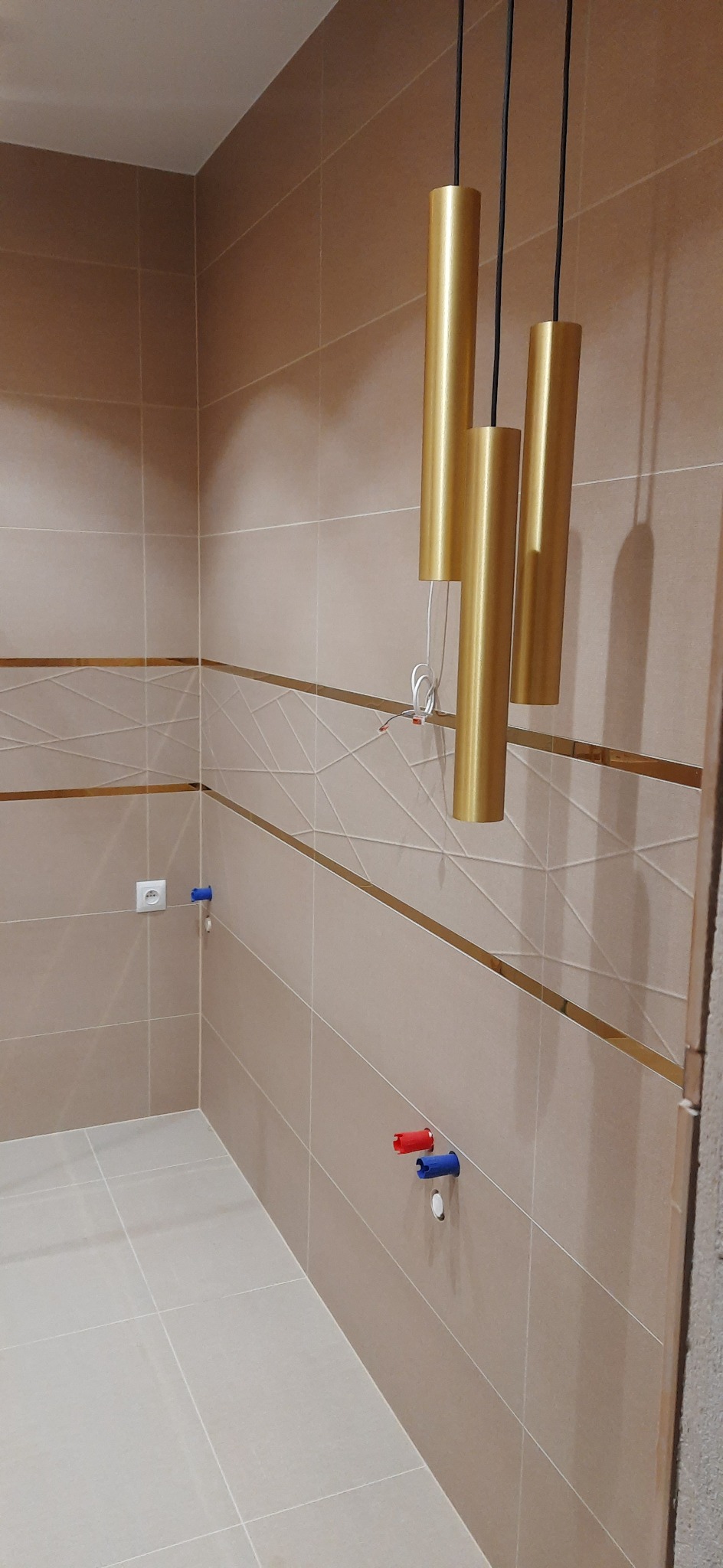 Wykończona łazienka z beżowymi płytkami z geometrycznym wzorem, złotymi, cylindrycznymi lampami wiszącymi i wyprowadzonymi instalacjami wodnymi z zaślepkami w kolorze czerwonym i niebieskim.