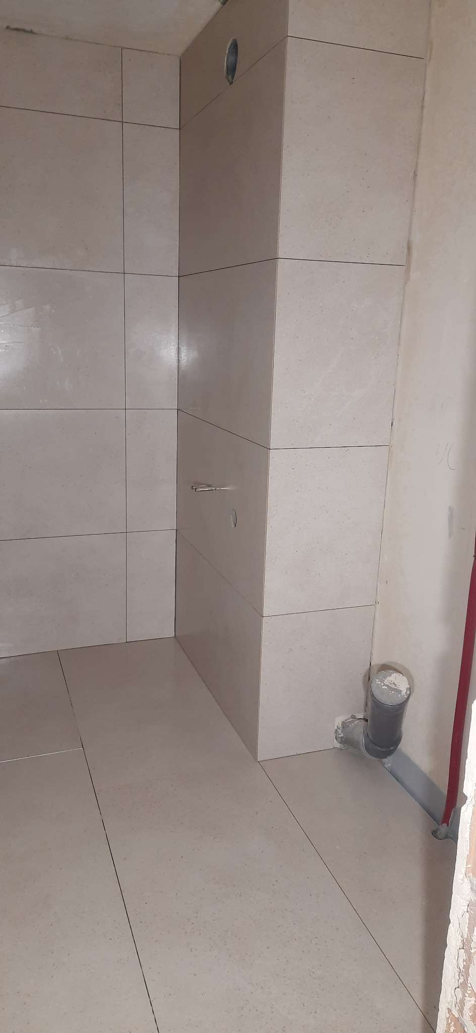 Fragment wykończonej łazienki z beżowymi płytkami ceramicznymi na ścianach i podłodze; widoczny odpływ kanalizacyjny przy ścianie oraz otwór w ścianie pod instalację.