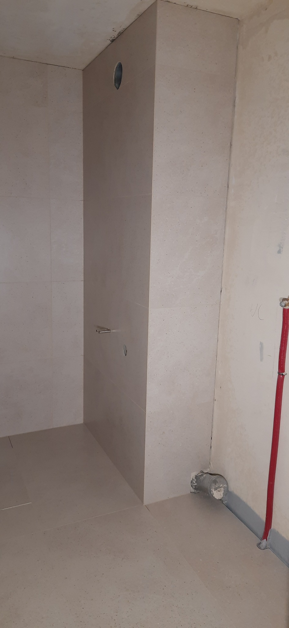 Surowy stan łazienki z beżowymi płytkami, widoczne rury hydrauliczne, otwór wentylacyjny i niedokończone ściany.