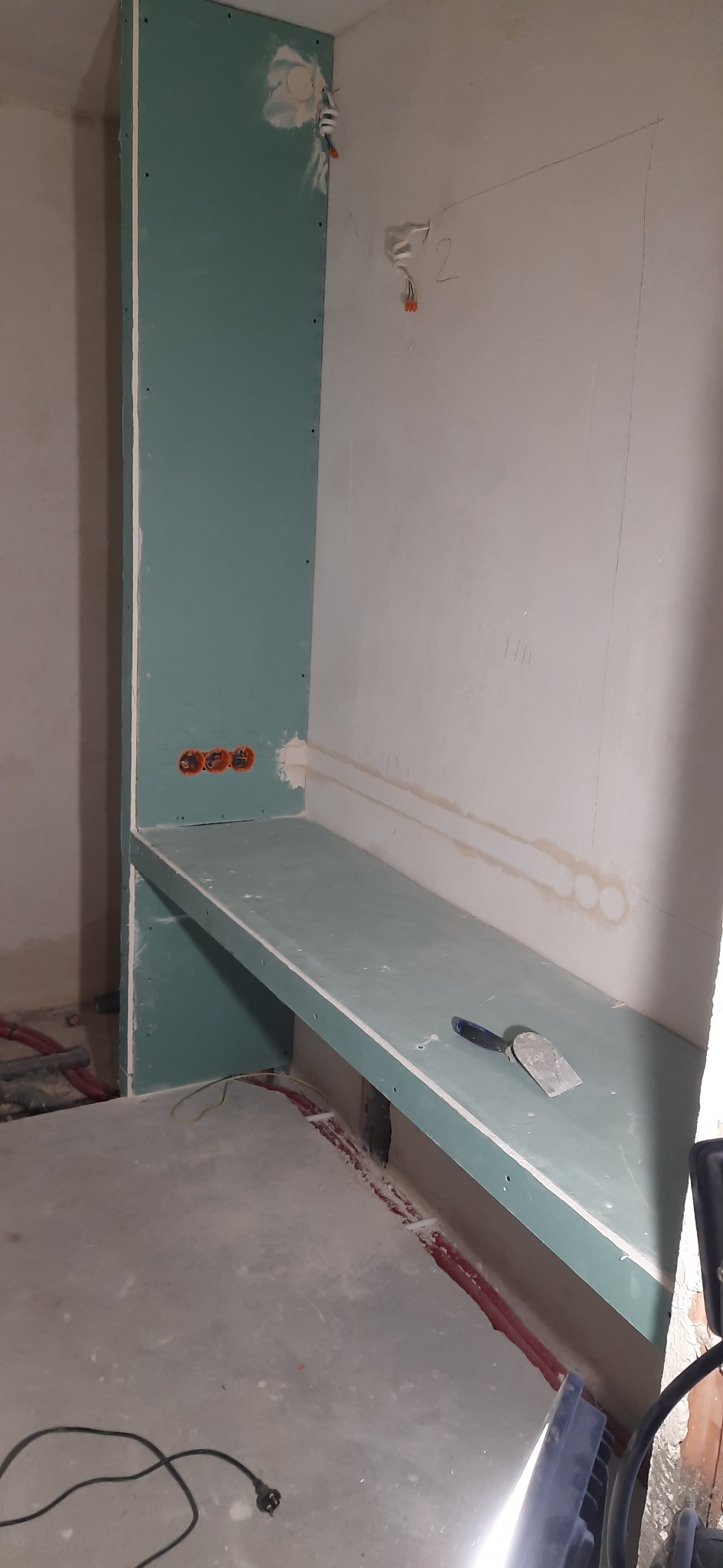 Surowy stan łazienki w trakcie remontu: zamontowana płyta gipsowo-kartonowa tworząca półkę, widoczne instalacje elektryczne i hydrauliczne w ścianach, szpachelka na półce, ślady po szpachlowaniu...