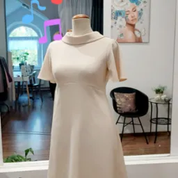 Kremowa sukienka z krótkim rękawem i stójką, prezentowana na drewnianym manekinie w pracowni krawieckiej, z widocznym odbiciem wnętrza w lustrze.