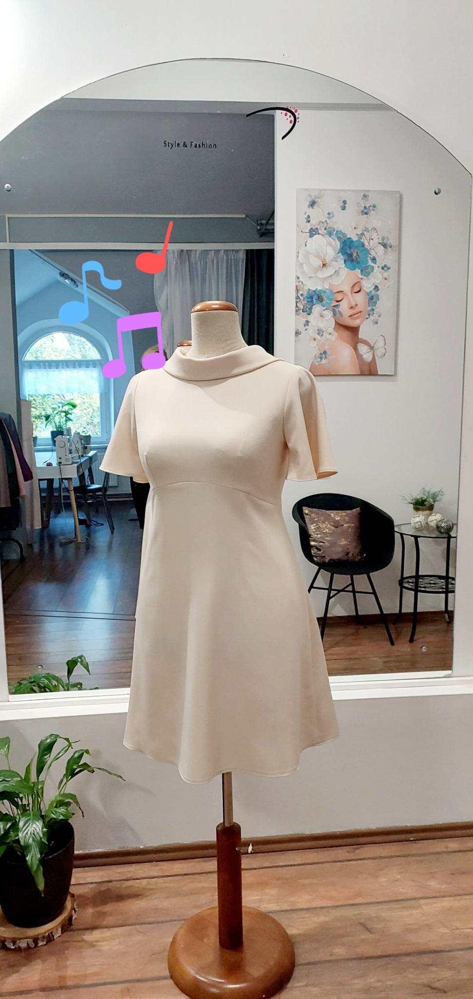 Kremowa sukienka z krótkim rękawem i stójką, prezentowana na drewnianym manekinie w pracowni krawieckiej, z widocznym odbiciem wnętrza w lustrze.
