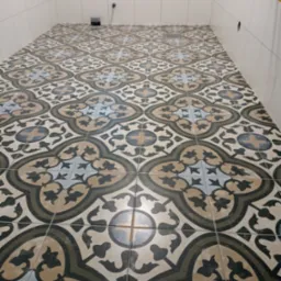 Wzorzysta, ułożona podłoga z płytek w łazience, widoczny odpływ liniowy i fragment ściany z białych płytek.