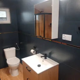 Nowoczesna łazienka z ciemnymi płytkami, drewnianą szafką pod umywalką z czarną armaturą, toaletą i lustrem z oświetleniem LED, w odbiciu widoczna kabina prysznicowa.