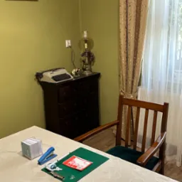 Biurko z zieloną podkładką, na której leżą formularze KP, zszywacz, notatnik, lampka na komodzie z maszyną do pisania w tle