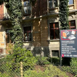 Tablica reklamowa z napisem 'BIURO RACHUNKOWE' i informacjami o usługach, umieszczona przed budynkiem z balkonem i pnącą się roślinnością.