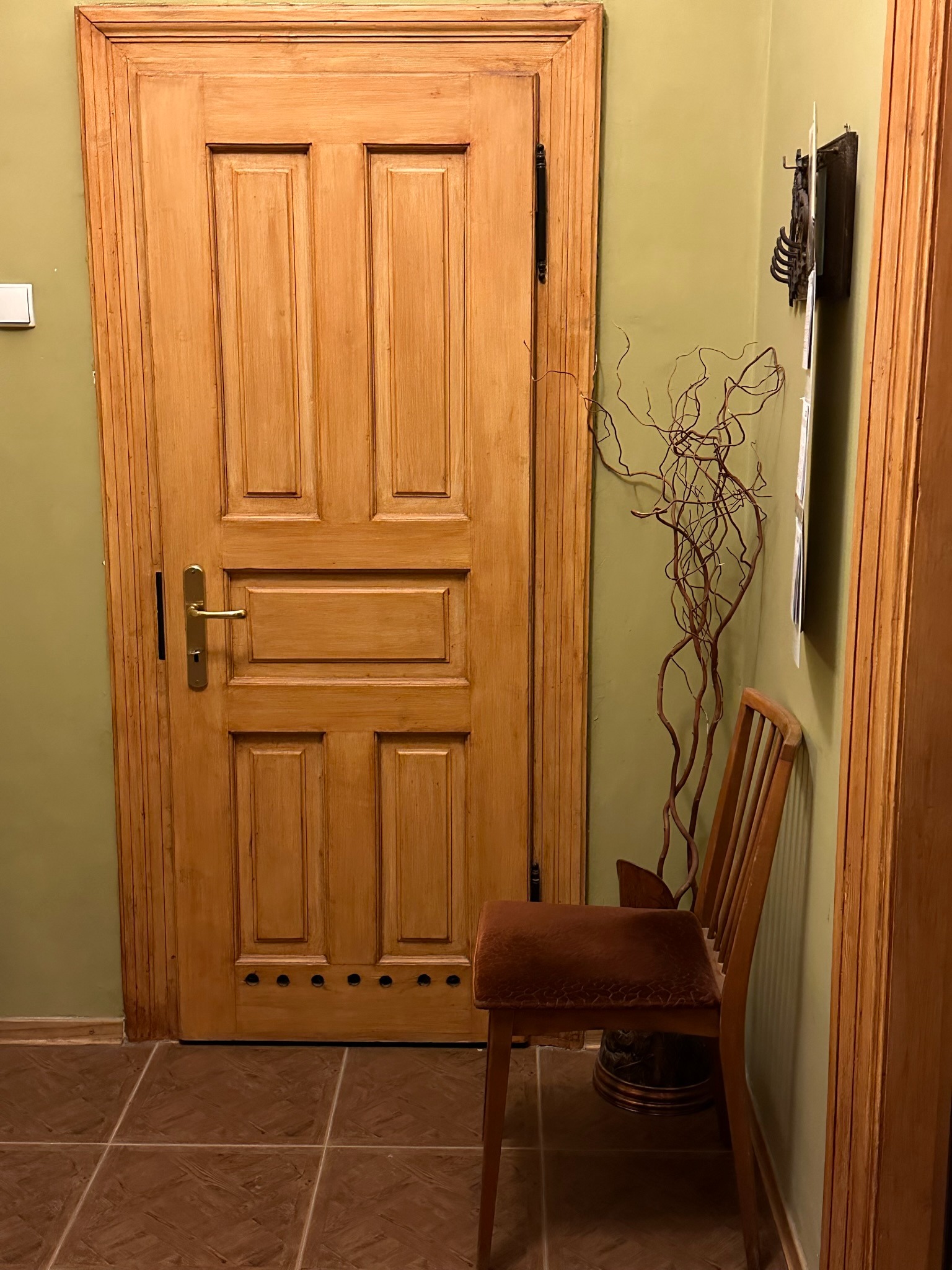 Drewniane drzwi wewnętrzne z metalową klamką, brązowa krzesło z welurowym siedziskiem, dekoracyjne gałęzie w wazonie, wieszak na ścianie w odcieniu zieleni, brązowe płytki podłogowe.