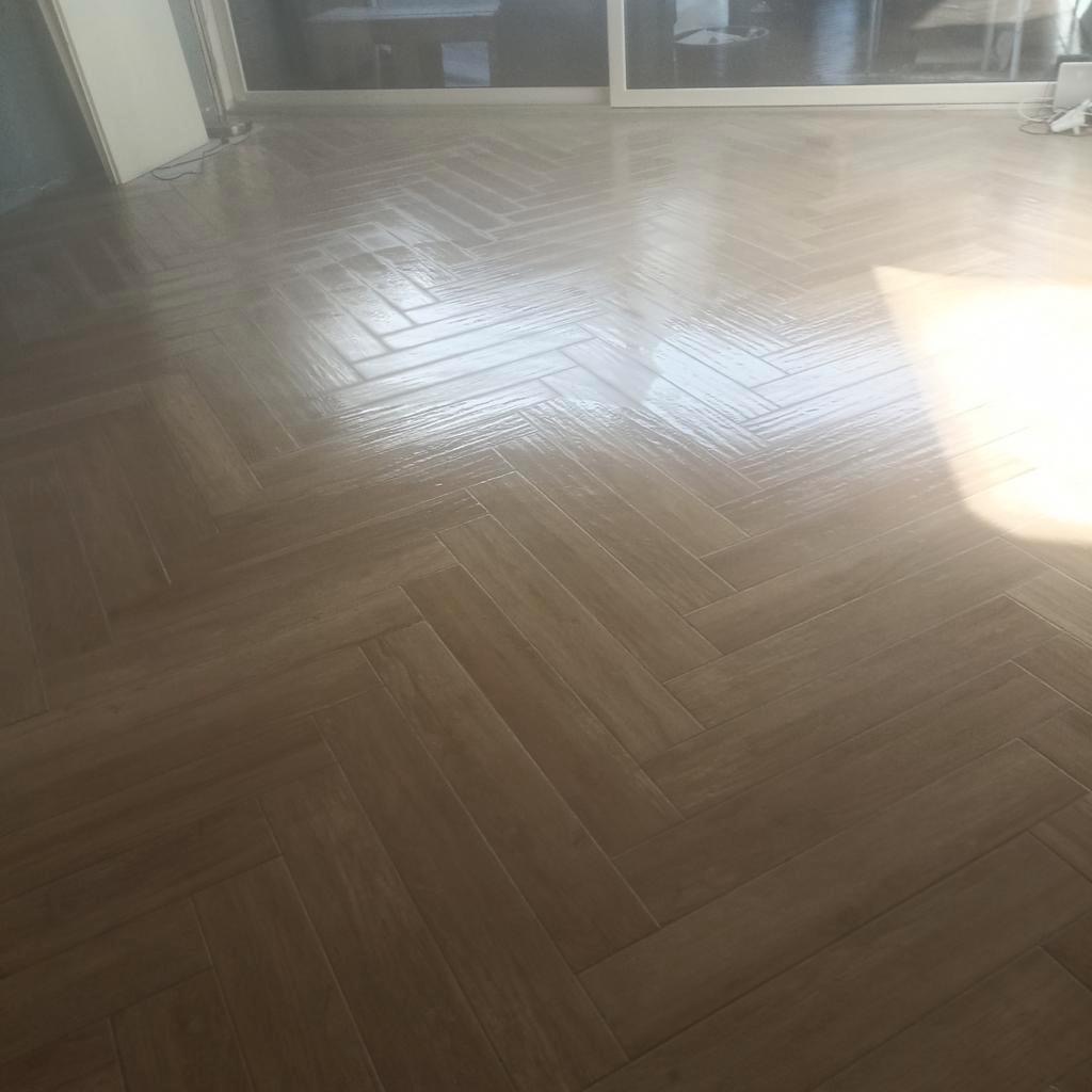 Ułożona w jodełkę, błyszcząca podłoga z płytek imitujących drewno w łazience, odbijająca światło dzienne.