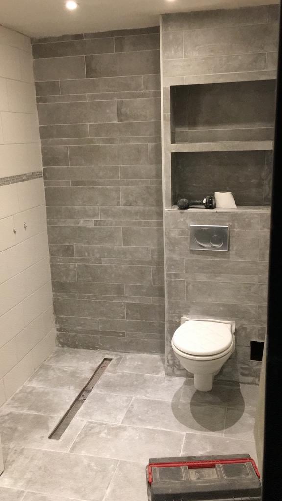 Świeżo wykafelkowana łazienka z szarymi płytkami imitującymi kamień, liniowy odpływ w podłodze, zabudowana półka ścienna z narzędziami i pojemnikiem na papier toaletowy, podwieszana toaleta...