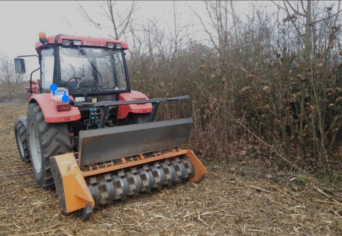 Czerwony traktor z zamontowanym frezem mulczującym na polu z resztkami roślinnymi i krzewami w tle.