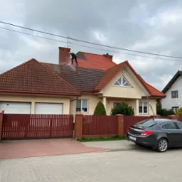 Pracownik w kombinezonie i kasku czyści czerwoną dach&oacute;wkę na dwuspadowym dachu domu jednorodzinnego za pomocą myjki ciśnieniowej, widoczny samoch&oacute;d osobowy i ogrodzenie.