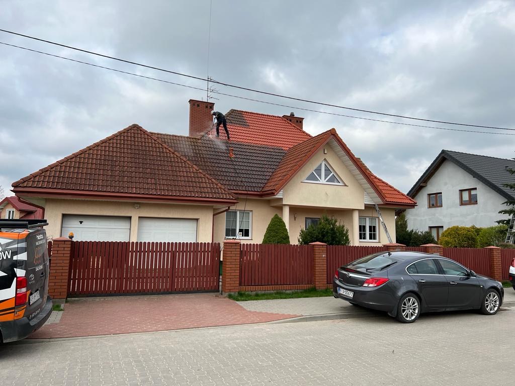 Pracownik w kombinezonie i kasku czyści czerwoną dachówkę na dwuspadowym dachu domu jednorodzinnego za pomocą myjki ciśnieniowej, widoczny samochód osobowy i ogrodzenie.