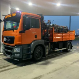 Pomarańczowa ciężarówka MAN z dźwigiem i ładunkiem rur stalowych na skrzyni, zaparkowana na stacji paliw pod zadaszeniem, widoczny numer telefonu na szybie czołowej.