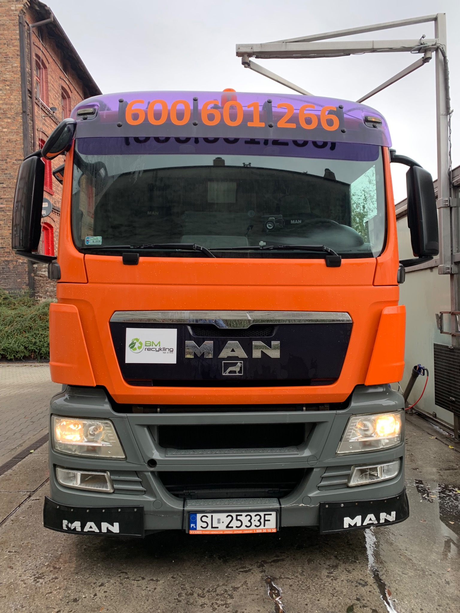 Pomarańczowa ciężarówka MAN z numerem telefonu na szybie, logo 'BM Recykling' i tablicą rejestracyjną SL 2533F, stojąca na mokrej nawierzchni przed budynkiem z czerwonej cegły.