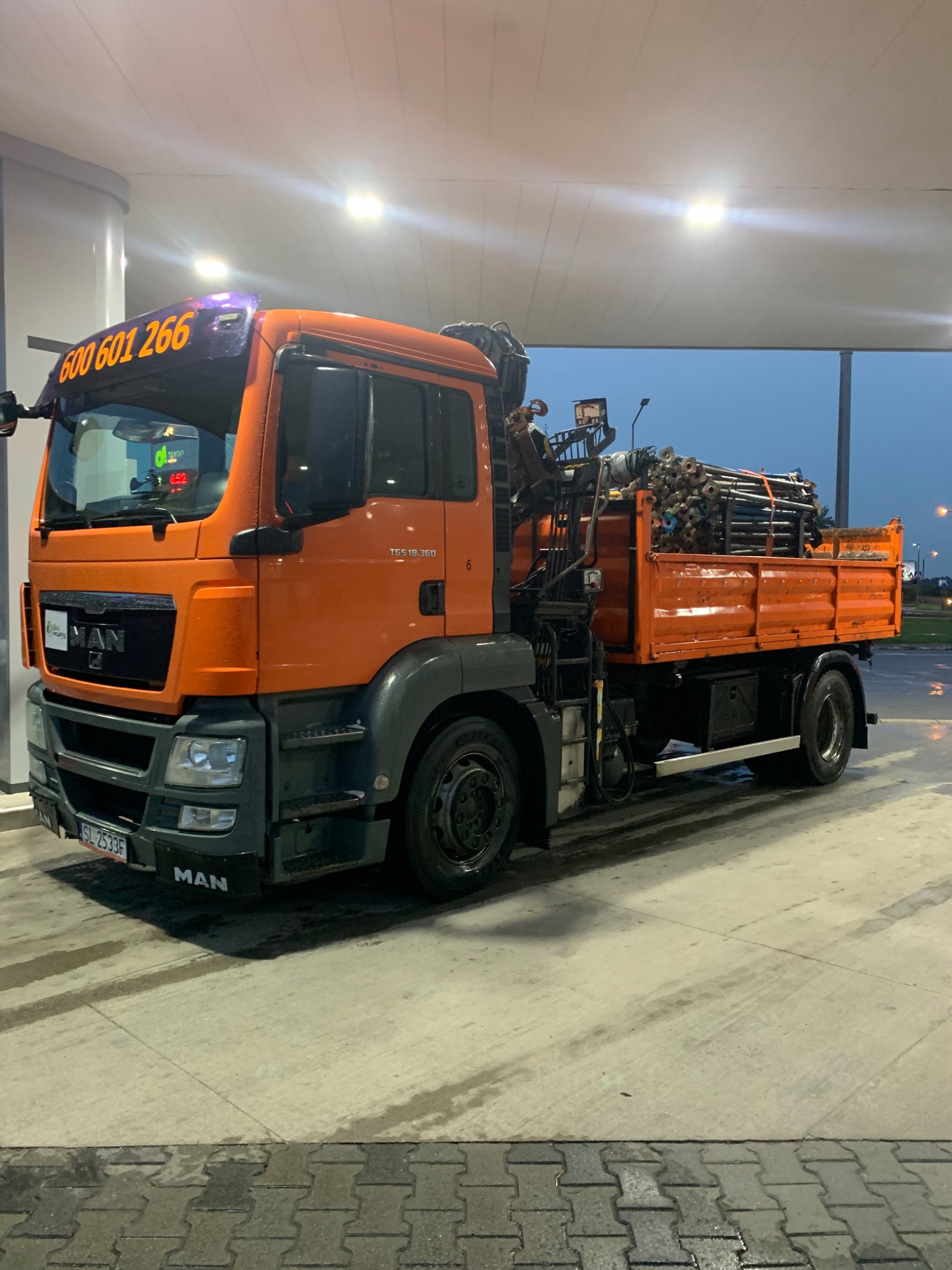 Pomarańczowa ciężarówka MAN z dźwigiem i ładunkiem rur stalowych na skrzyni, zaparkowana na stacji paliw pod zadaszeniem, widoczny numer telefonu na szybie czołowej.