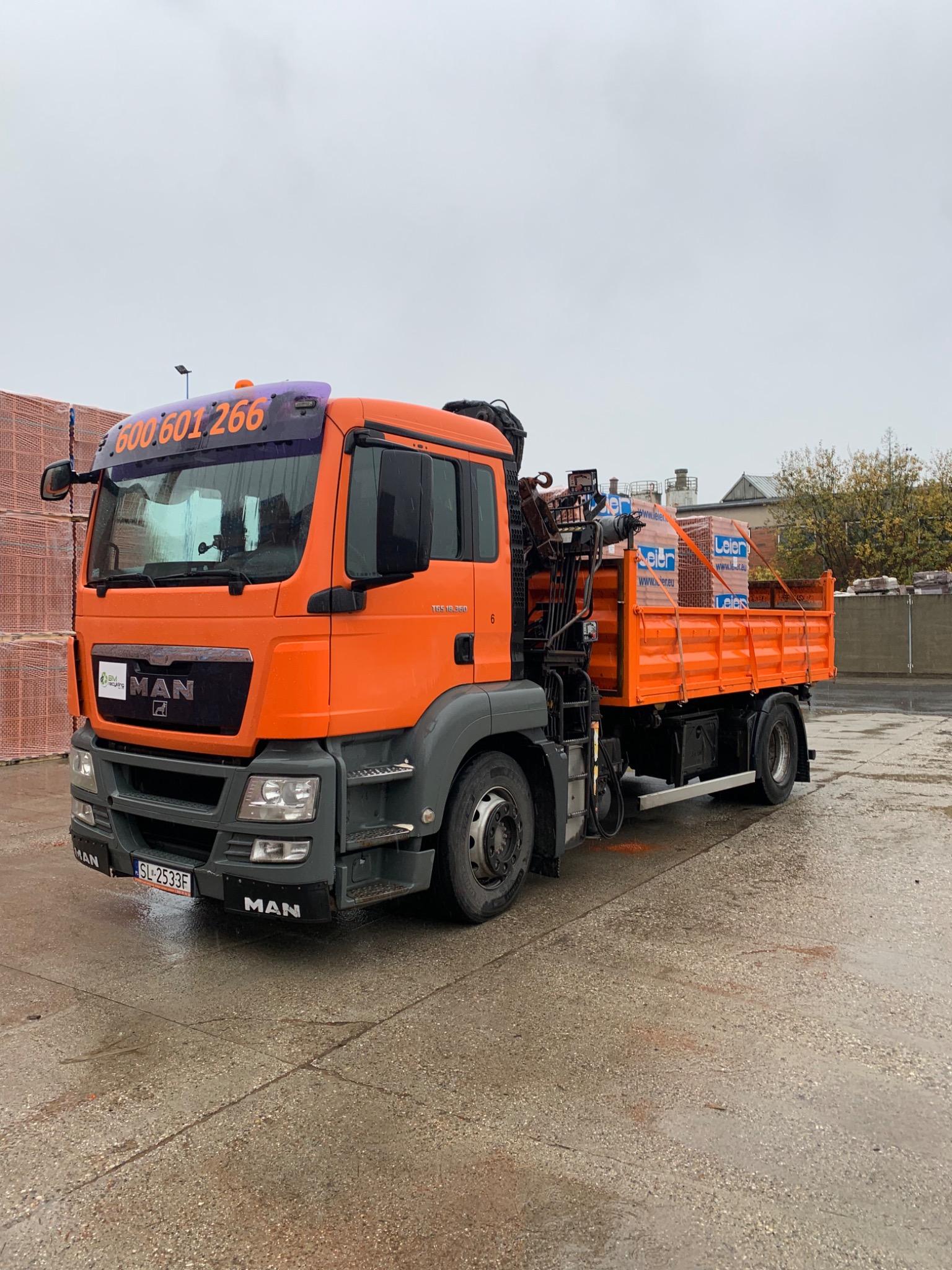 Pomarańczowy samochód ciężarowy MAN z dźwigiem, załadowany cegłami Leier, z numerem telefonu 600601266 na przedniej szybie, stojący na mokrym placu.