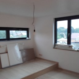 ABRemonty - Pusty pokój z dwiema oknami o czarnych ramach, jasną podłogą i niepodłączoną żarówką zwisającą z sufitu, przygotowany do prac wykończeniowych.