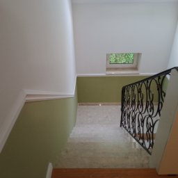 ABRemonty - Klatka schodowa po odświeżeniu: białe ściany z dekoracyjną listwą, oliwkowa ściana z małym oknem, marmurowe schody i czarna, ozdobna balustrada.