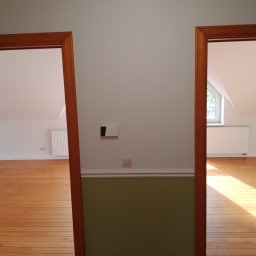 ABRemonty - Widok na dwa pokoje przez drewniane ościeżnice, z parkietem i grzejnikami pod skosami dachu, ściana pomalowana do połowy na oliwkowy kolor.