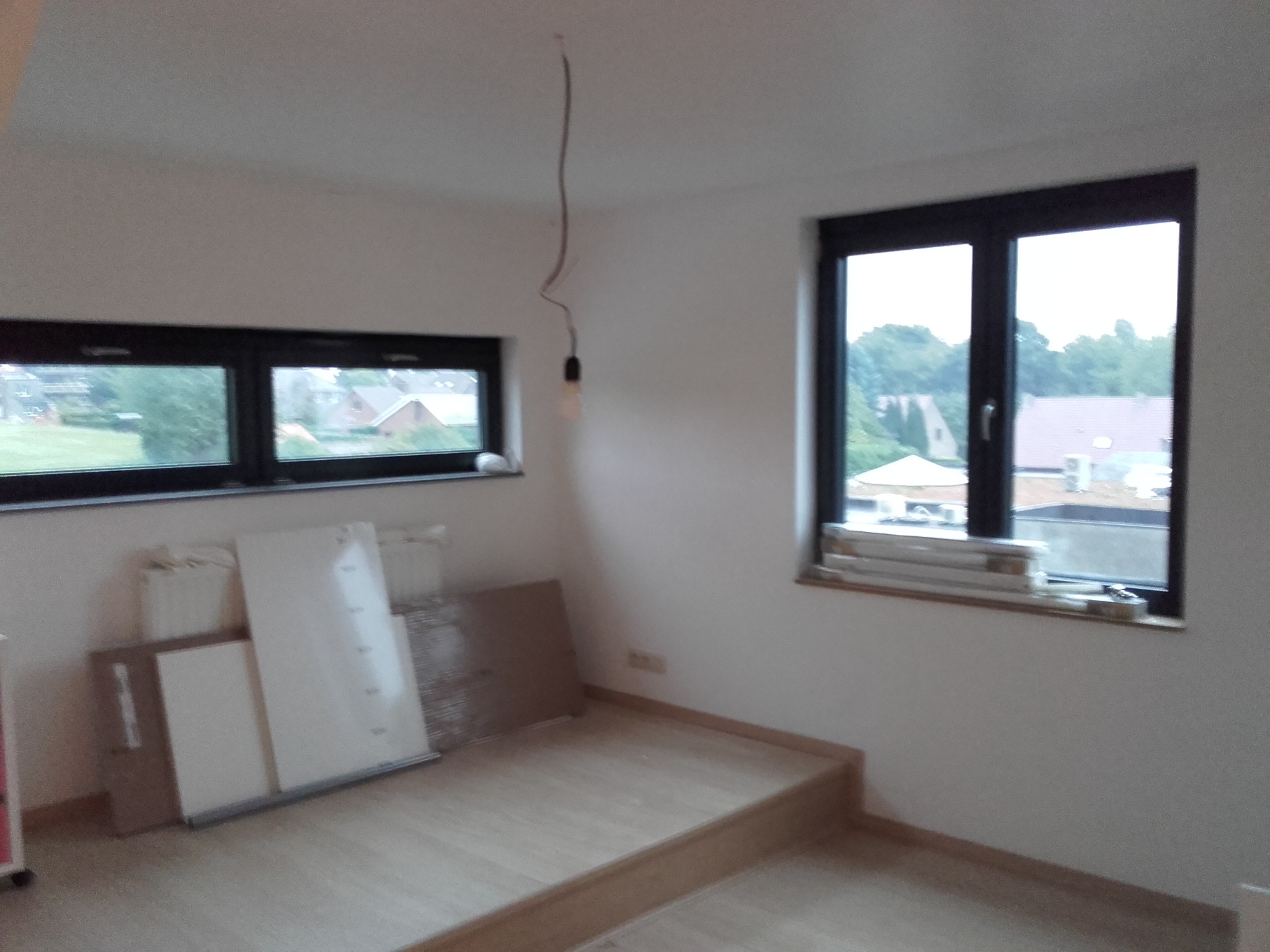 Pusty pokój z dwiema oknami o czarnych ramach, jasną podłogą i niepodłączoną żarówką zwisającą z sufitu, przygotowany do prac wykończeniowych.