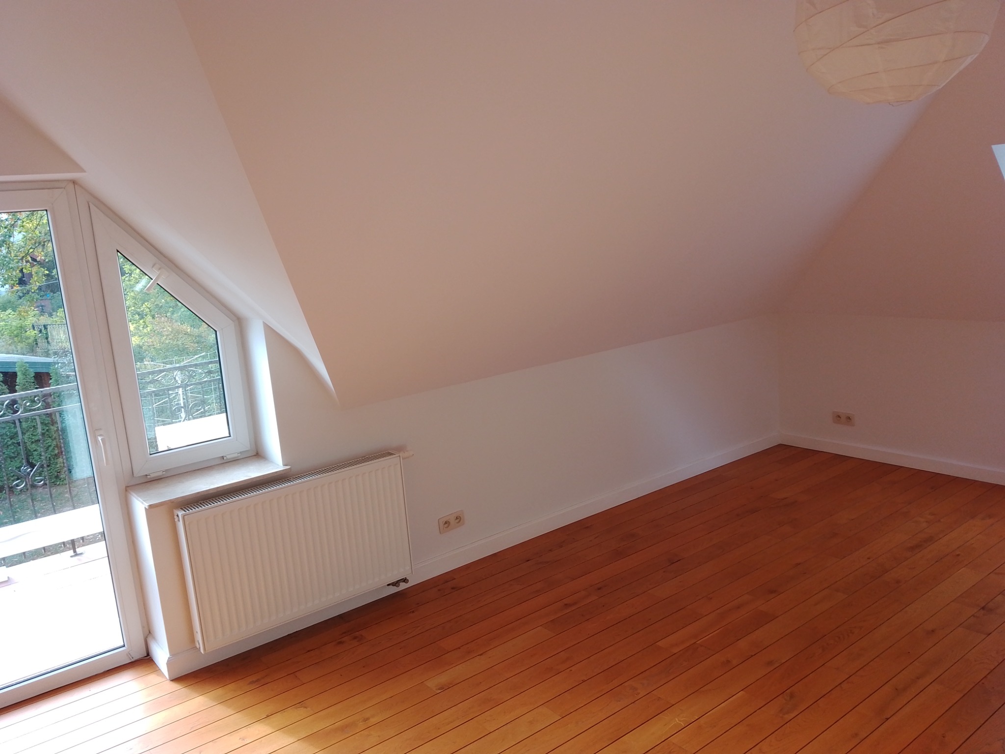 Poddasze po remoncie: świeżo pomalowane białe ściany i ułożony parkiet z naturalnego drewna, widoczne gniazdka elektryczne, okno balkonowe z widokiem na zieleń i biały grzejnik.