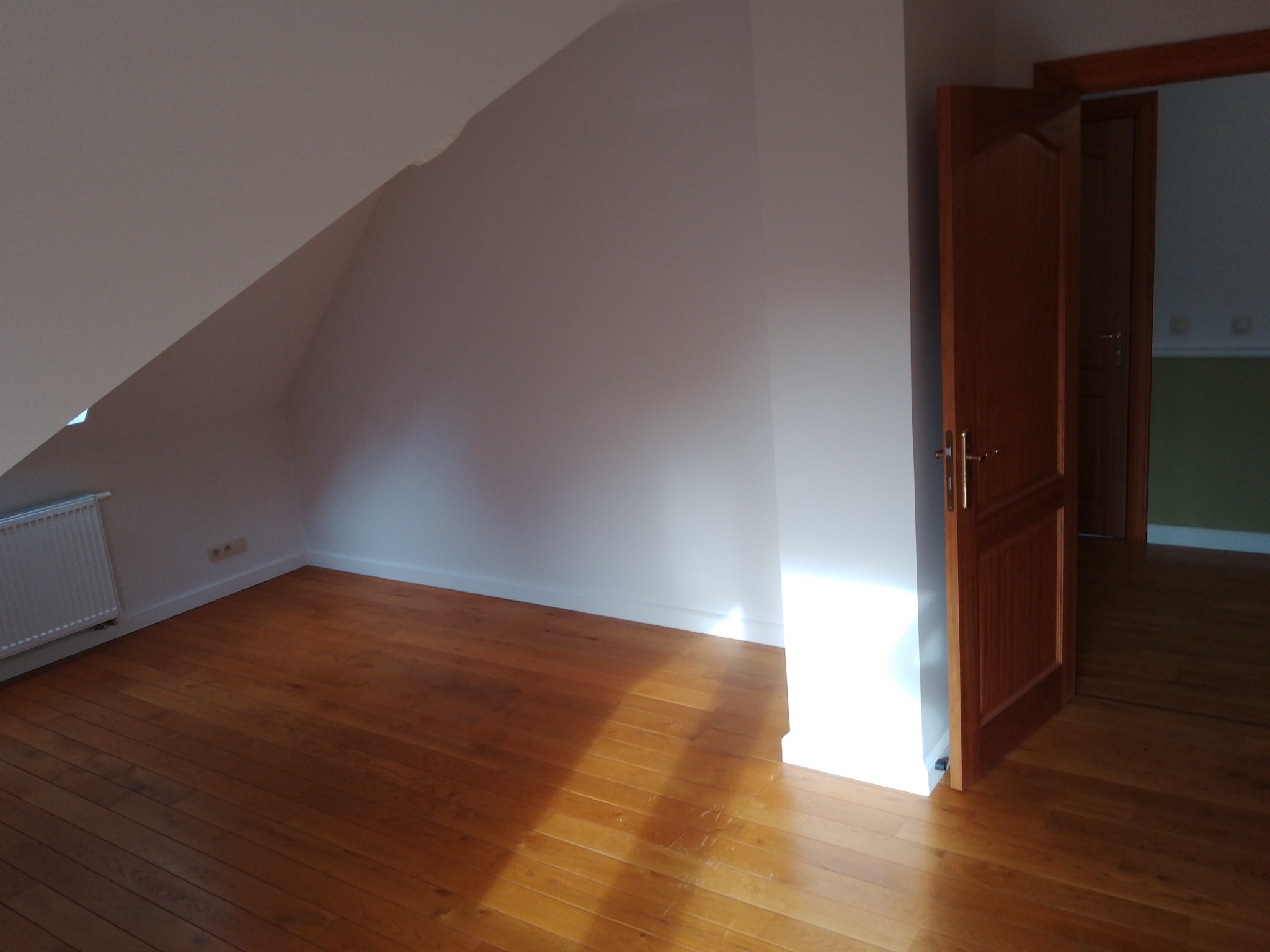 Poddasze z drewnianą podłogą, świeżo pomalowanymi białymi ścianami i otwartymi drewnianymi drzwiami, przez które widać fragment sąsiedniego pokoju.