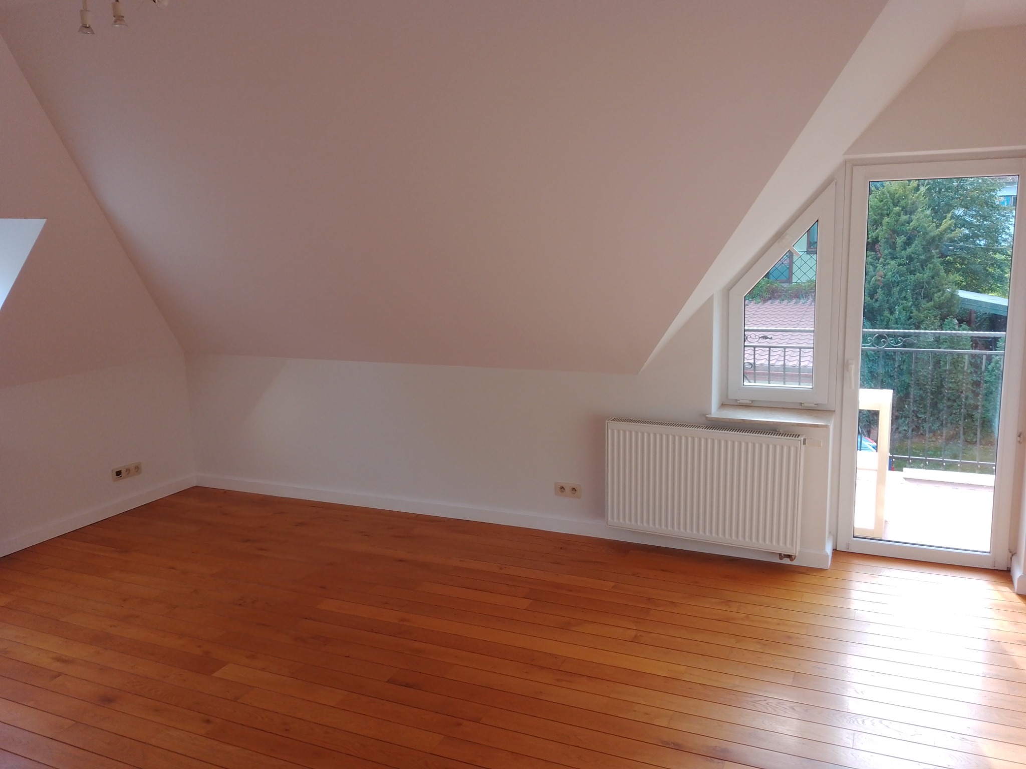 Puste poddasze z drewnianą podłogą, białymi ścianami i grzejnikiem pod oknem balkonowym z widokiem na zieleń.