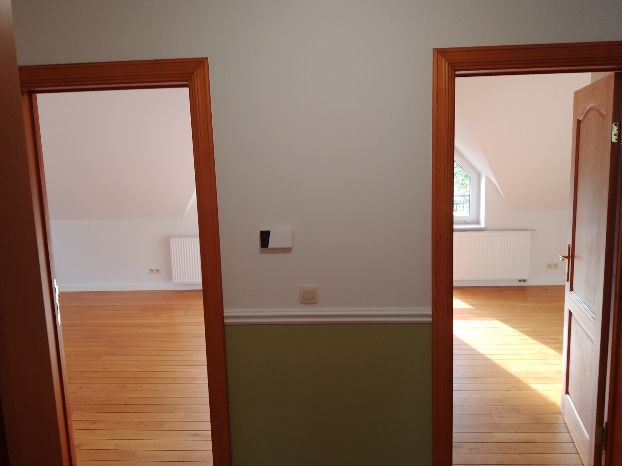 Widok na dwa pokoje przez drewniane ościeżnice, z parkietem i grzejnikami pod skosami dachu, ściana pomalowana do połowy na oliwkowy kolor.