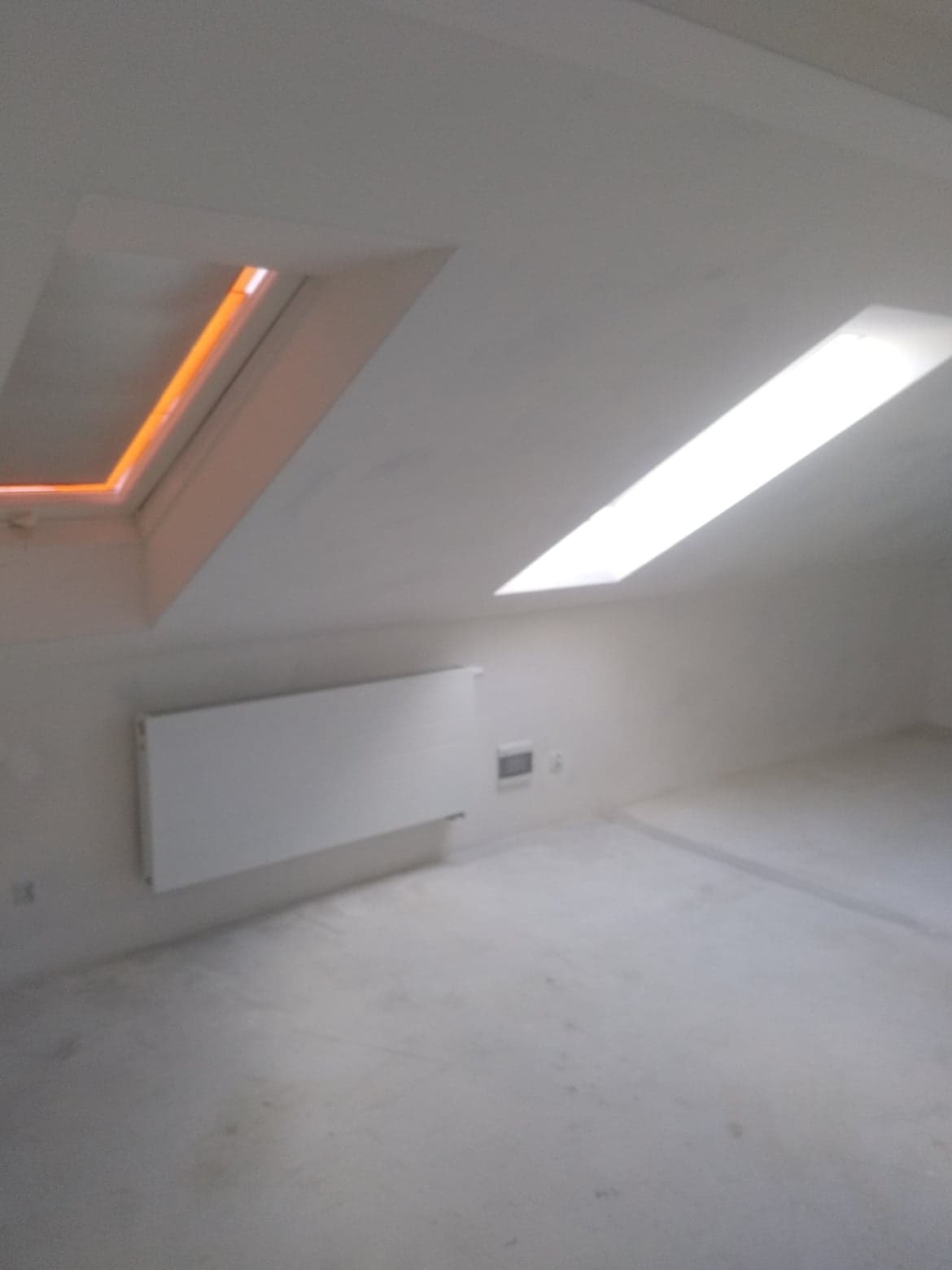 Poddasze po nałożeniu gładzi, widoczne dwa okna dachowe - jedno z pomarańczową poświatą, drugie z białym światłem dziennym, grzejnik na ścianie.