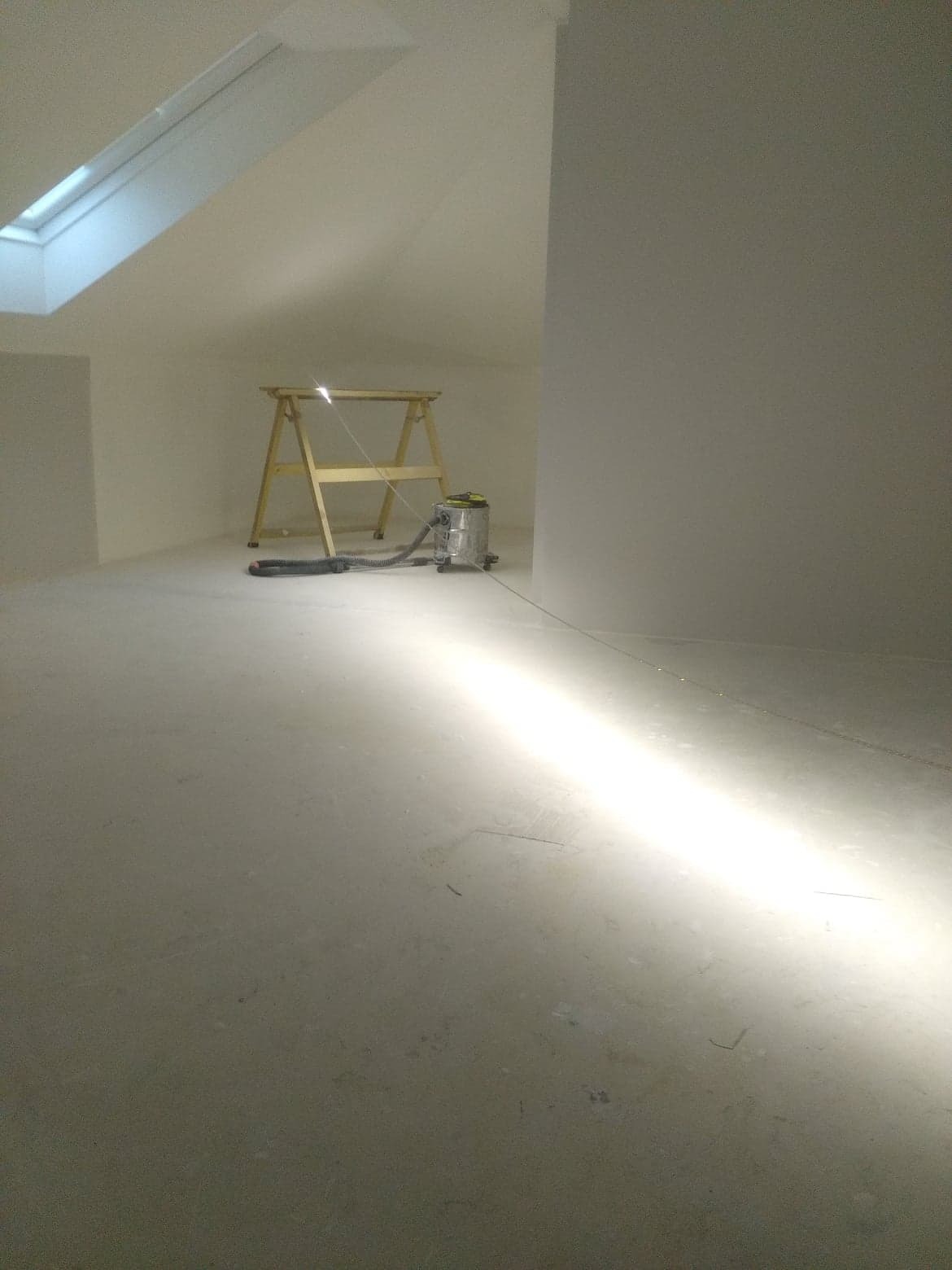 Poddasze po wylaniu posadzki, widoczny drewniany kozioł malarski i odkurzacz przemysłowy w tle, oświetlone światłem z okna dachowego.