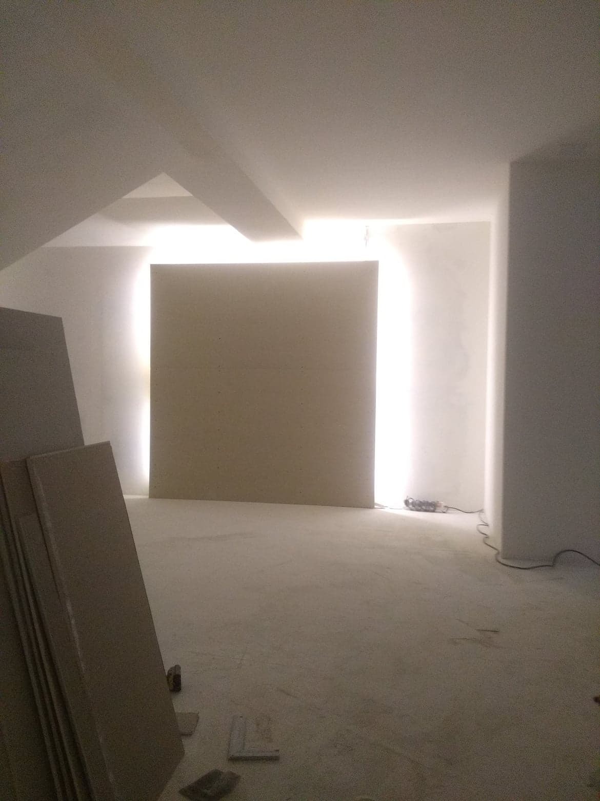 Wnętrze w trakcie prac wykończeniowych, widoczne płyty gipsowo-kartonowe oświetlone od tyłu, surowa podłoga i ściany przygotowane do dalszych prac.