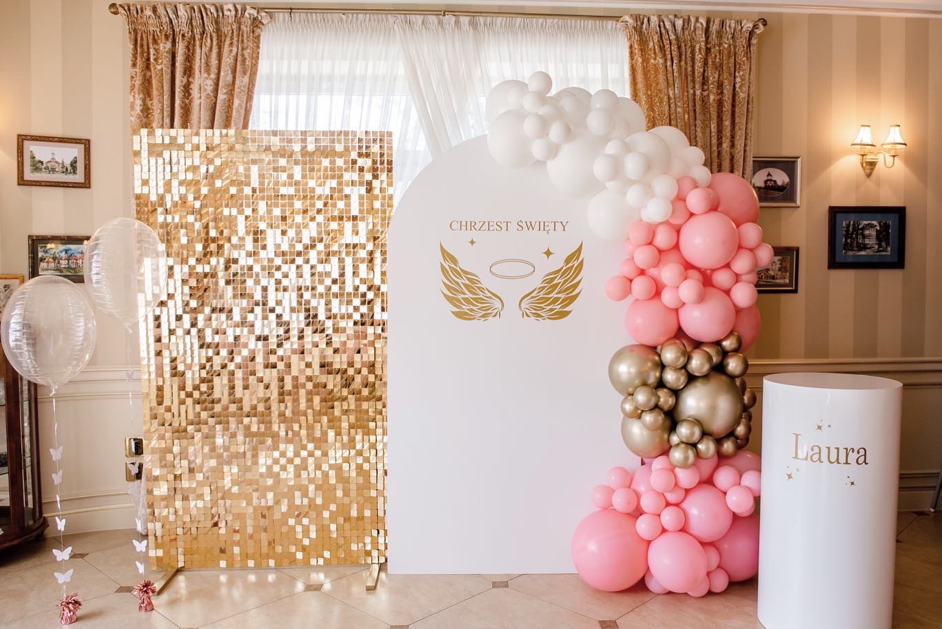 Dekoracja na Chrzest Święty z błyszczącą ścianką, balonami w kolorach białym, różowym i złotym oraz białym postumentem z imieniem Laura, umieszczona w eleganckim wnętrzu.